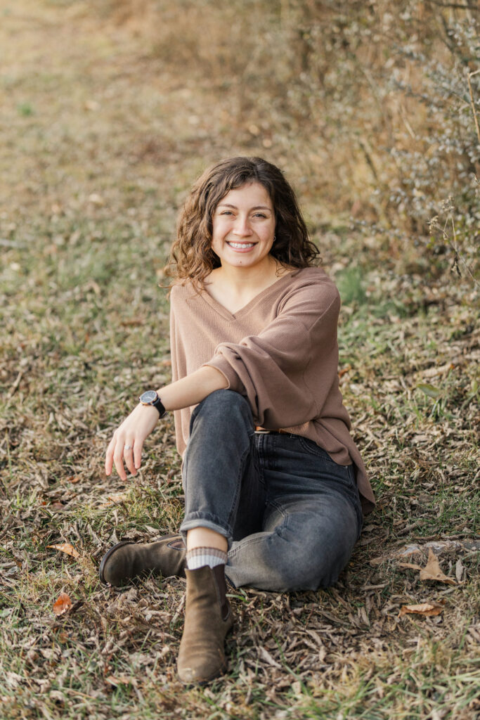 Woman sitting on the ground smiling wearing a mauve sweater.